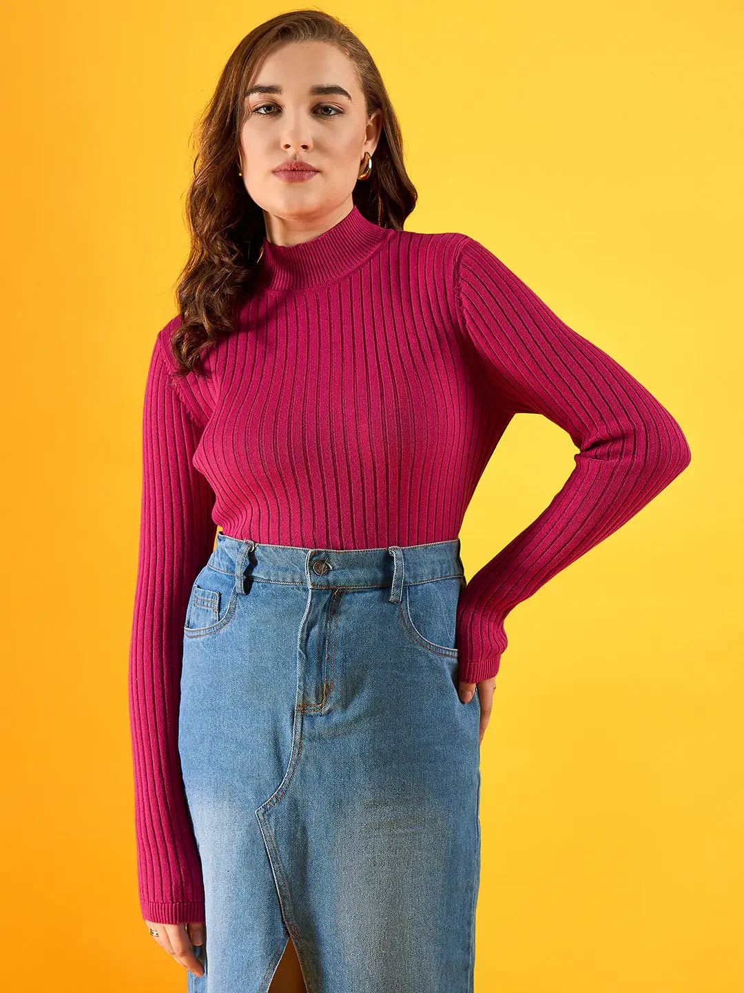 Woman in a 98degreenorth fuchsia ribbed high neck sweater and denim skirt stands against a yellow background.