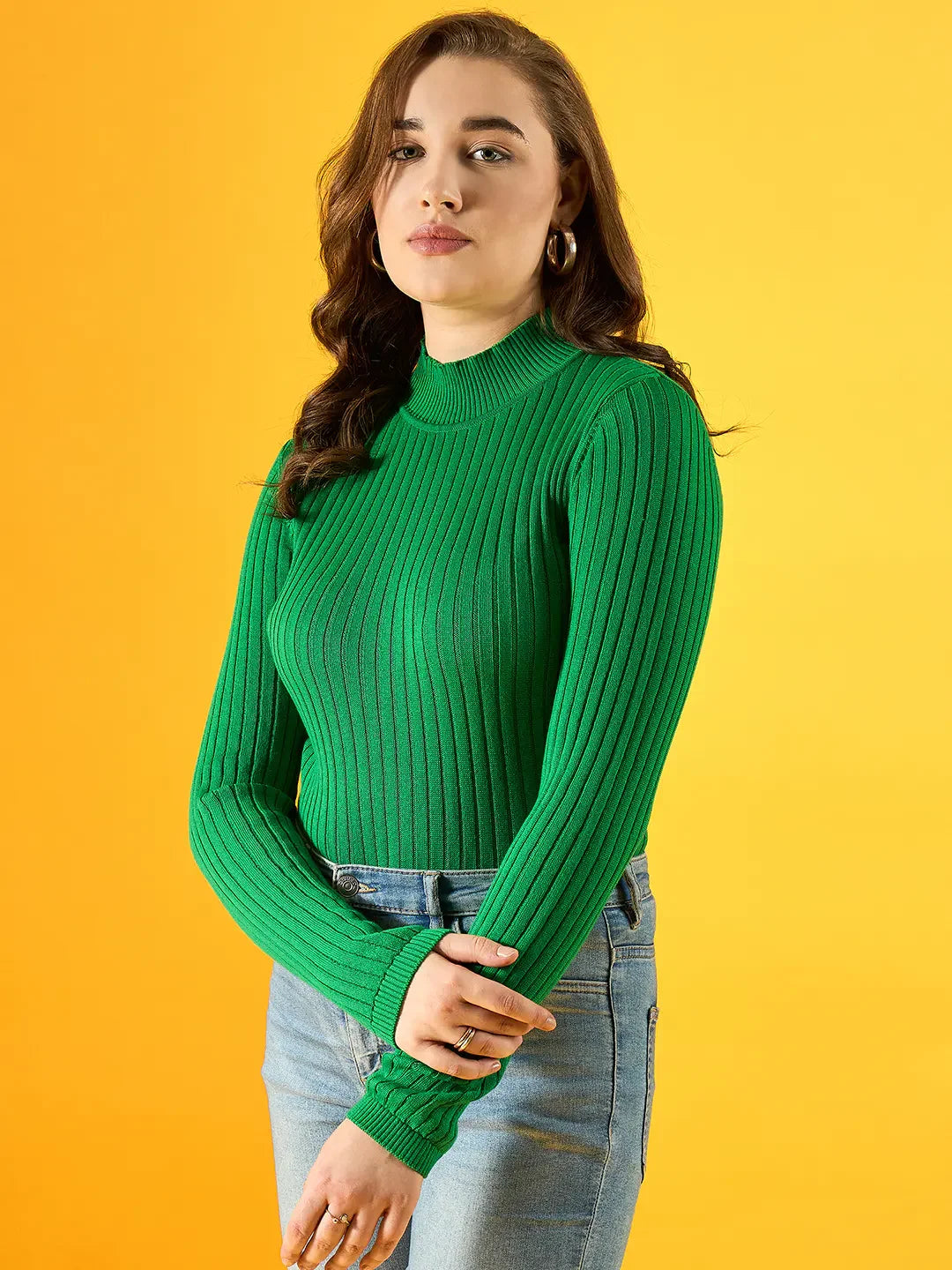 Woman wears 98degreenorth Women Ribbed High Neck Sweater–Green, paired with blue jeans, against a yellow background.
