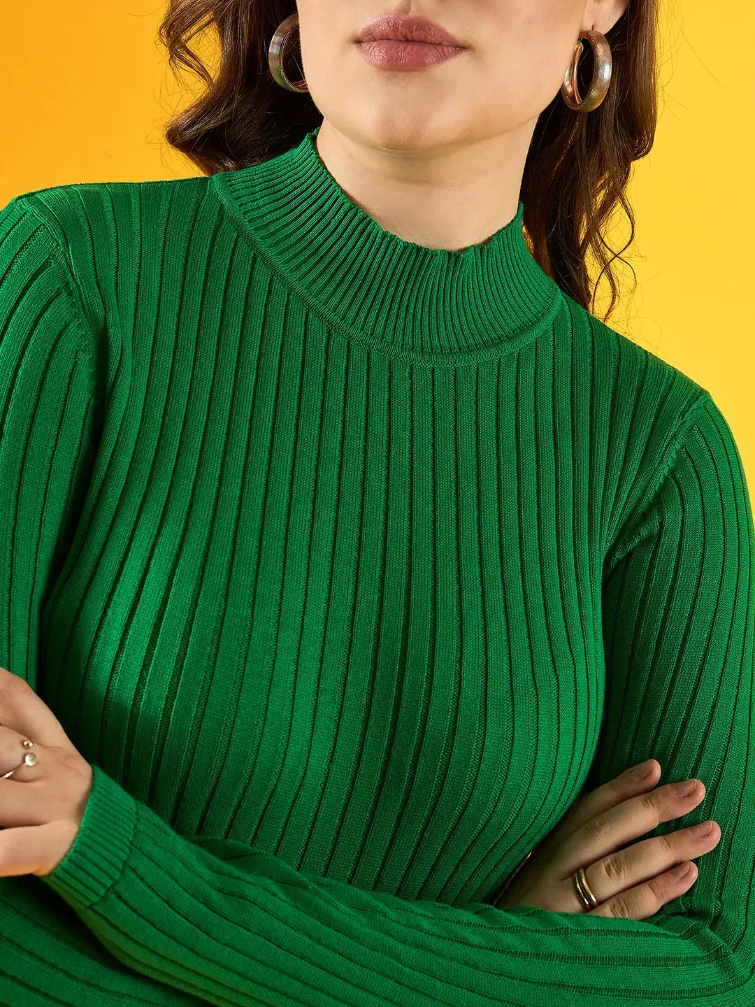 Woman wears 98degreenorth green ribbed high neck sweater, arms crossed, set before a yellow background.