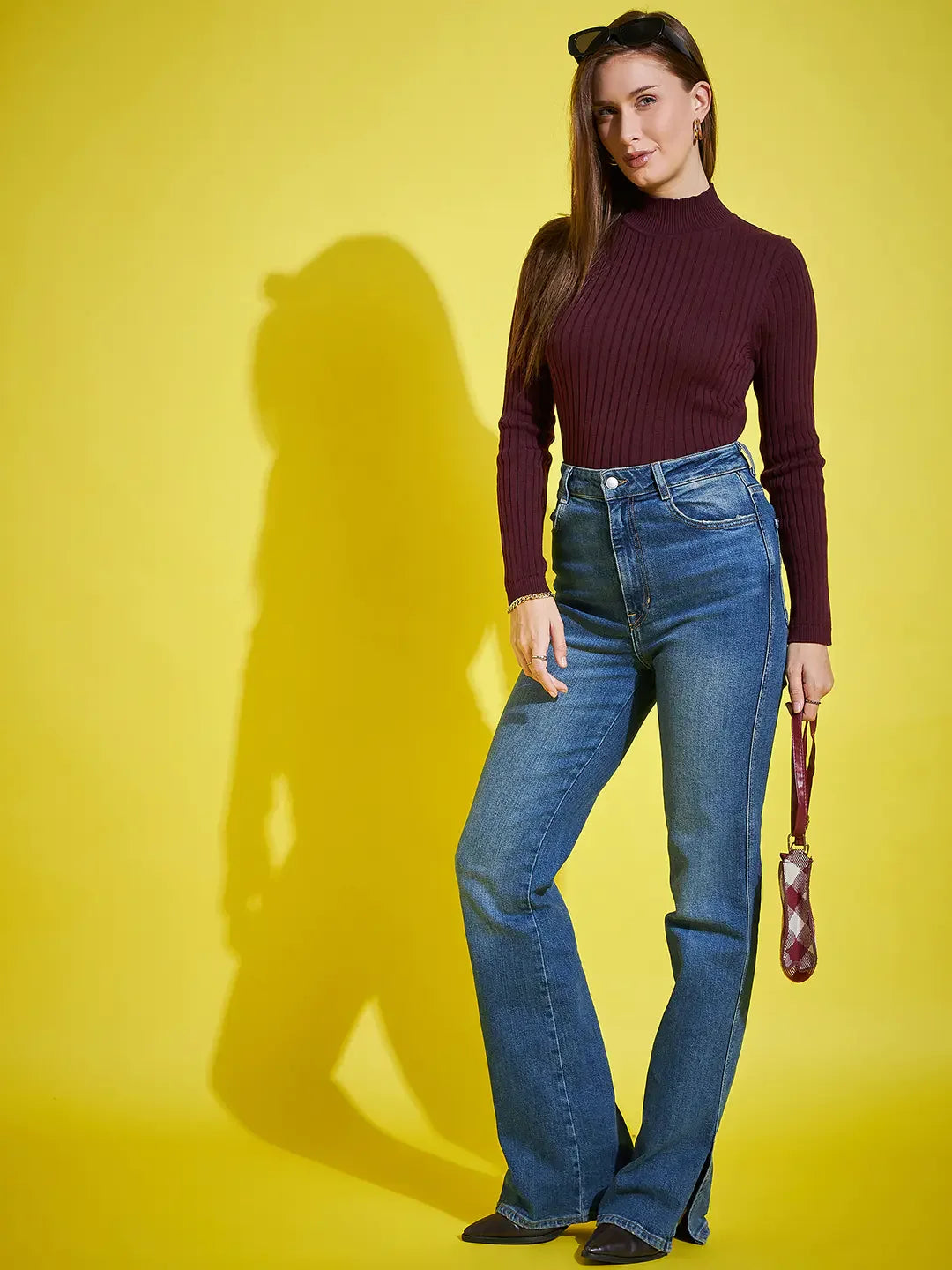Woman in a 98degreenorth Ribbed High Neck Wine Sweater stands against yellow, holding a handbag and sunglasses.