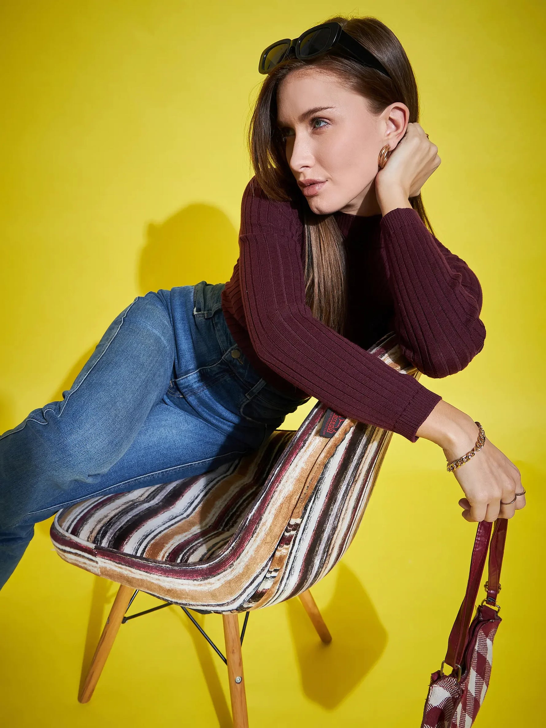Woman models 98degreenorth’s Wine Ribbed High Neck Sweater—soft winter pullover—paired with jeans on a striped chair.