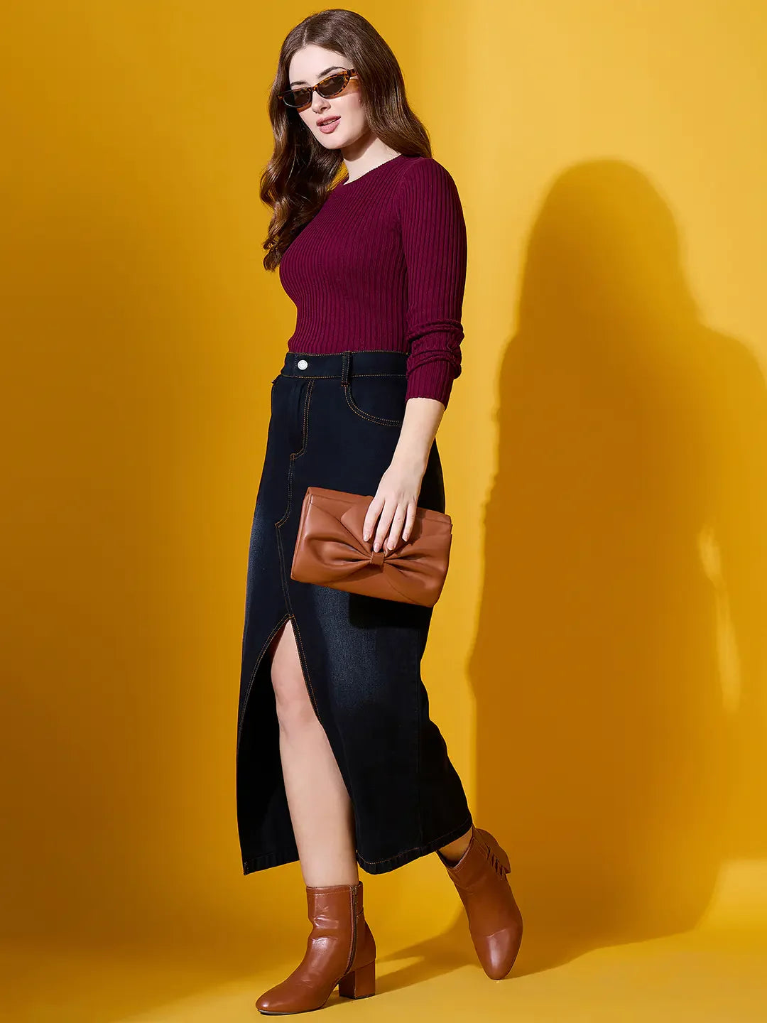 Woman in 98degreenorth Rosewood Ribbed Sweater, dark denim skirt, brown boots—chic winter style on yellow.