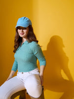 Woman in a blue cap and 98degreenorth Sea Green Ribbed Collar Neck Sweater sits on a stool against yellow backdrop.