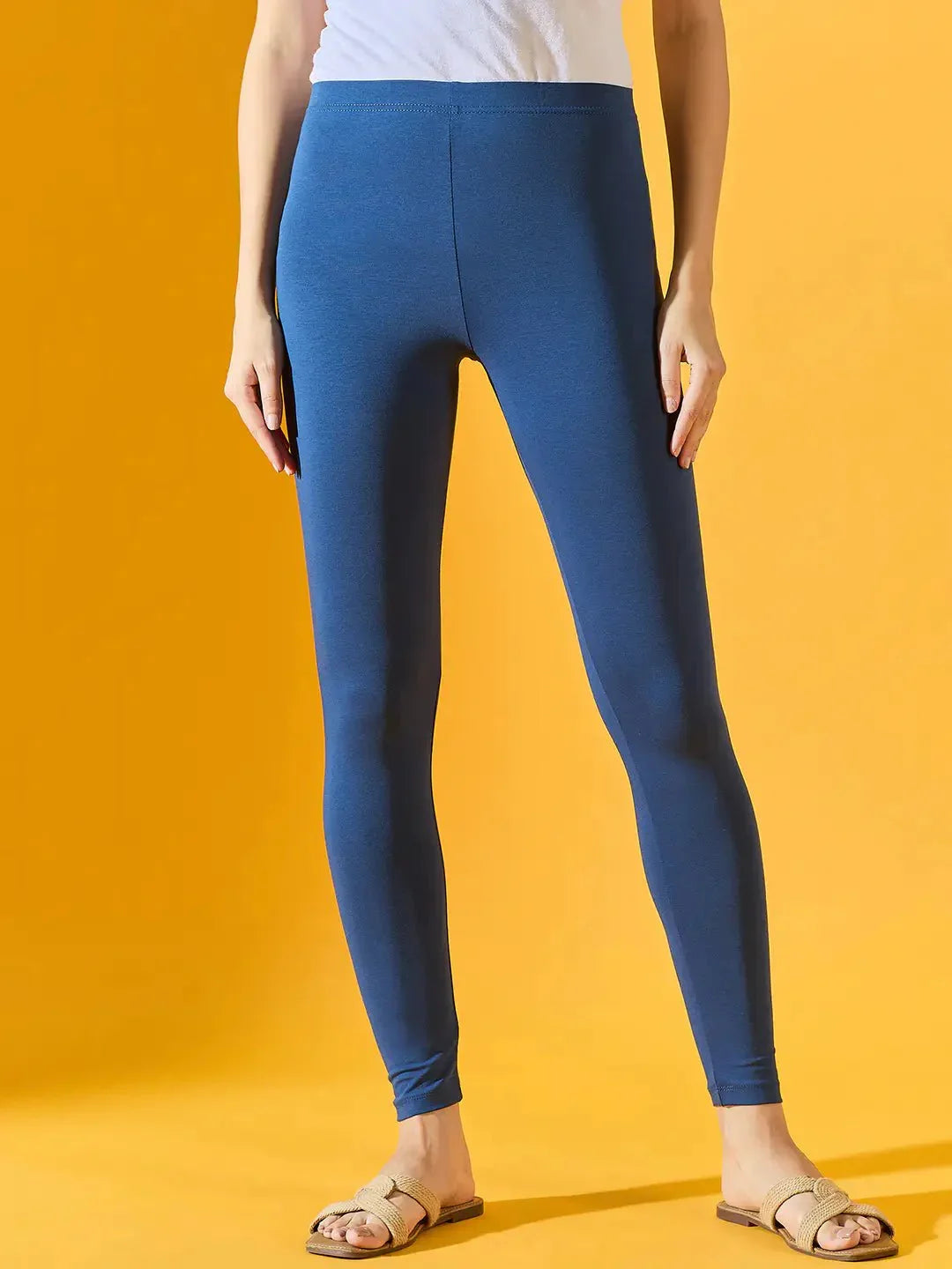 Model in 98degreenorth Women Teal Blue Ankle-Length Leggings, white top, and sandals poses by a yellow backdrop.