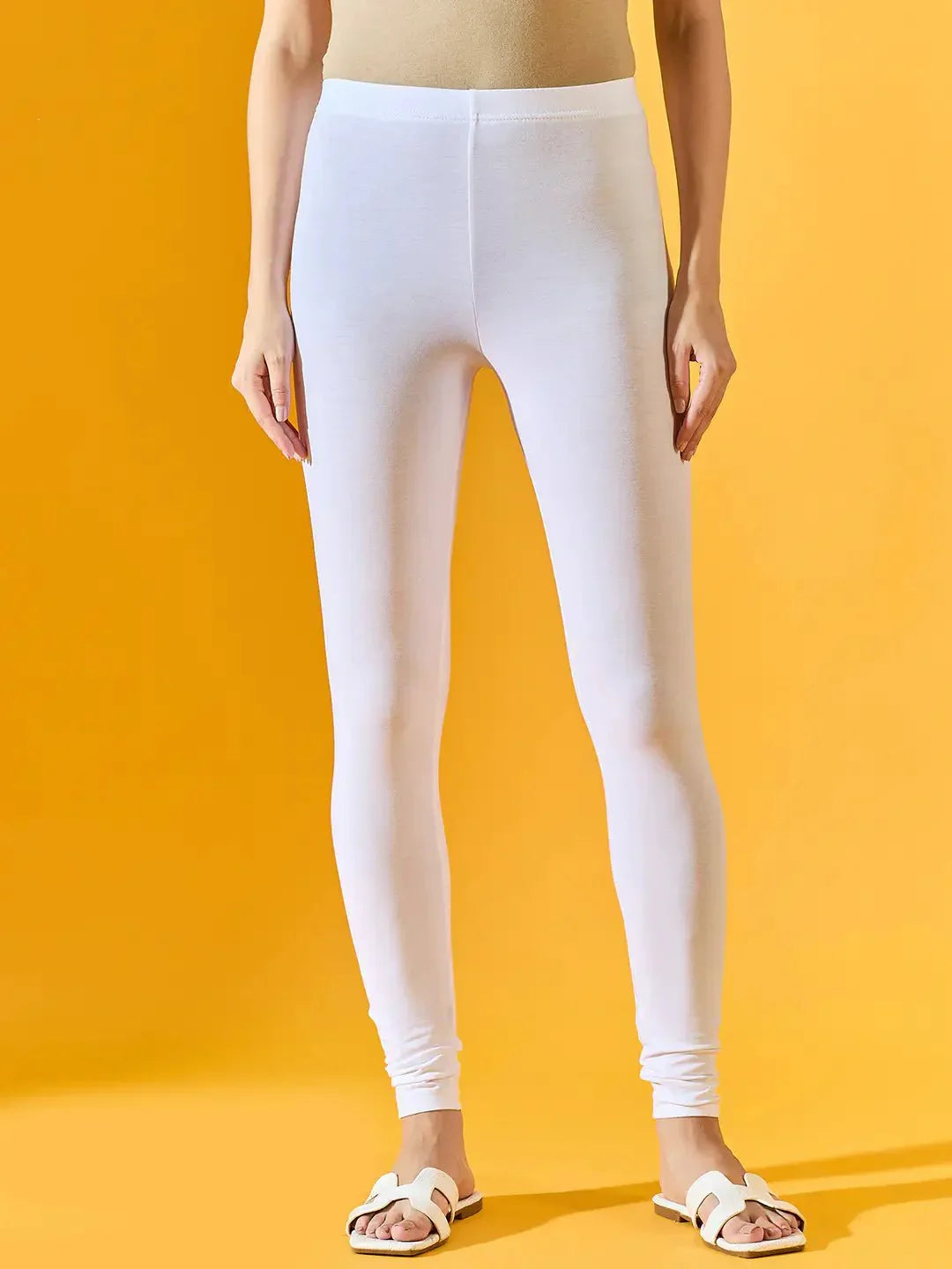 Only the lower half of the body is shown, featuring 98degreenorth’s Women White Ankle-Length Leggings against a yellow background. The model pairs them with a beige top and white sandals for a casual look.