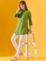 A woman wearing 98degreenorth Women White Ankle-Length Leggings stands against a yellow background, leaning on a white stool and touching her hair, paired with a green kurti and white sandals.
