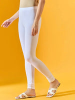 A person models the 98degreenorth Women White Ankle-Length Leggings, paired with a beige top and white sandals, standing against a solid yellow background. Only the lower body is shown.