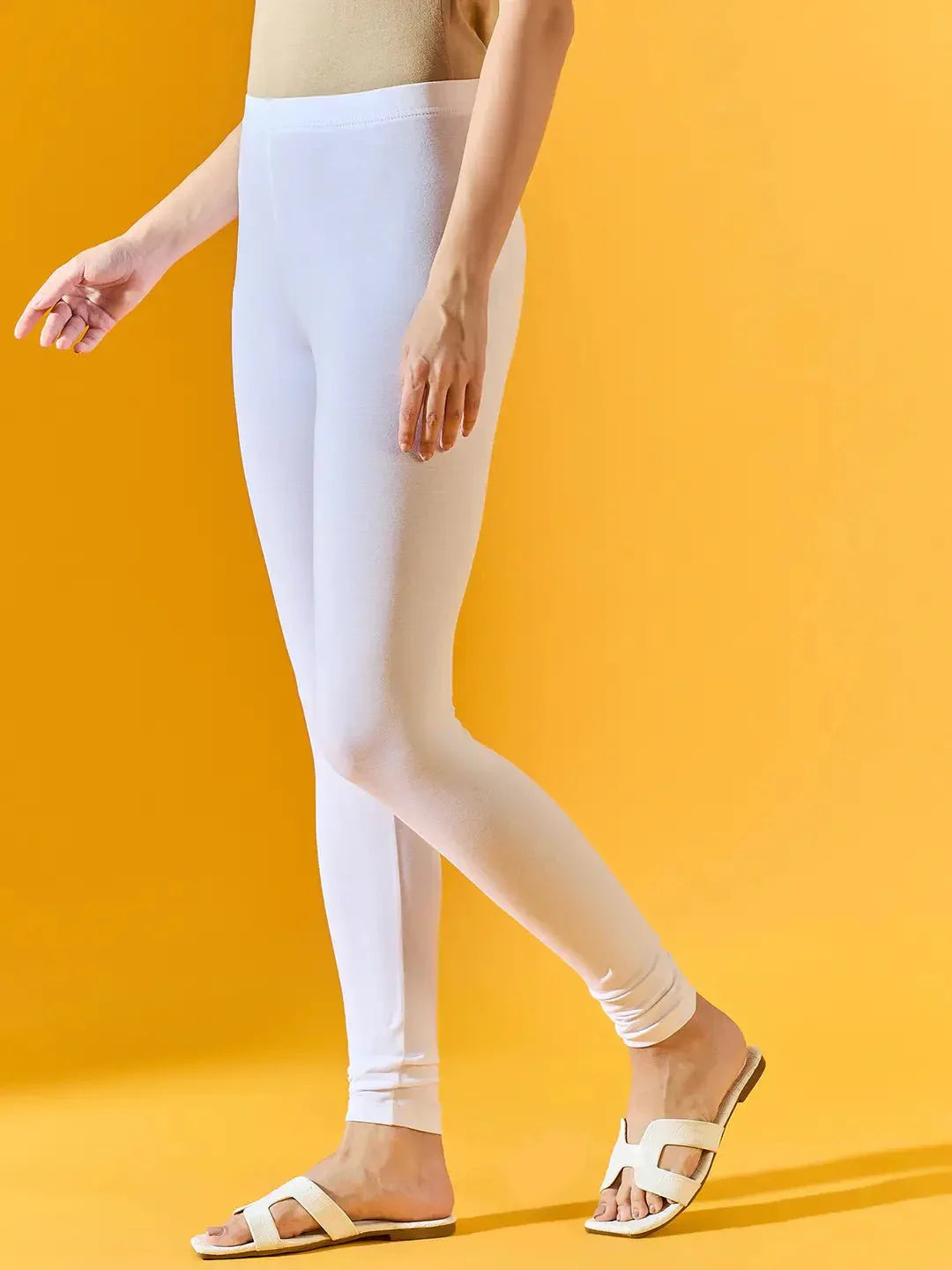 A person models the 98degreenorth Women White Ankle-Length Leggings, paired with a beige top and white sandals, standing against a solid yellow background. Only the lower body is shown.
