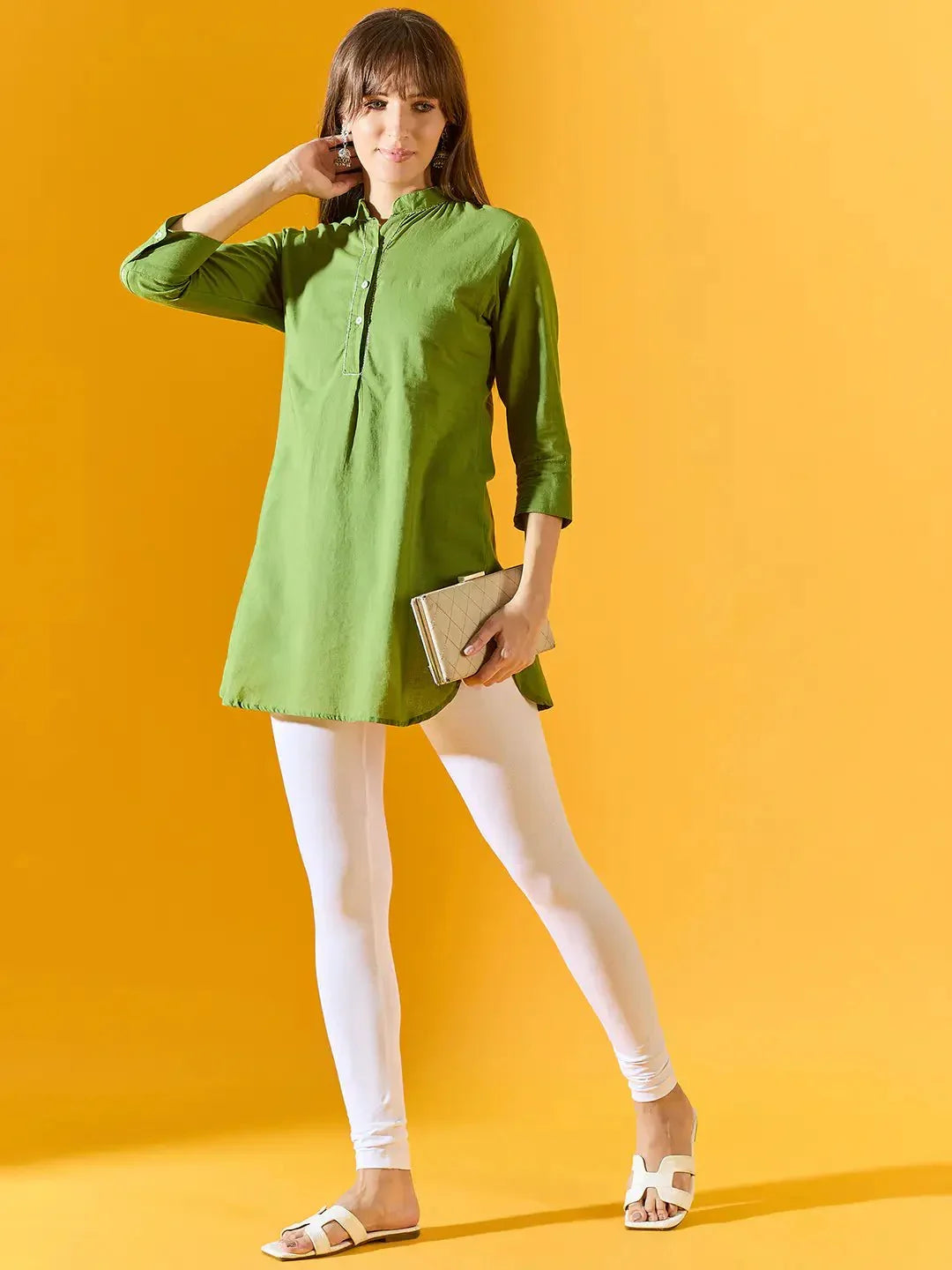 A woman stands against a yellow background wearing 98degreenorth Women White Ankle-Length Leggings, a green long-sleeve tunic, and white sandals. She holds a beige clutch and casually touches her hair.