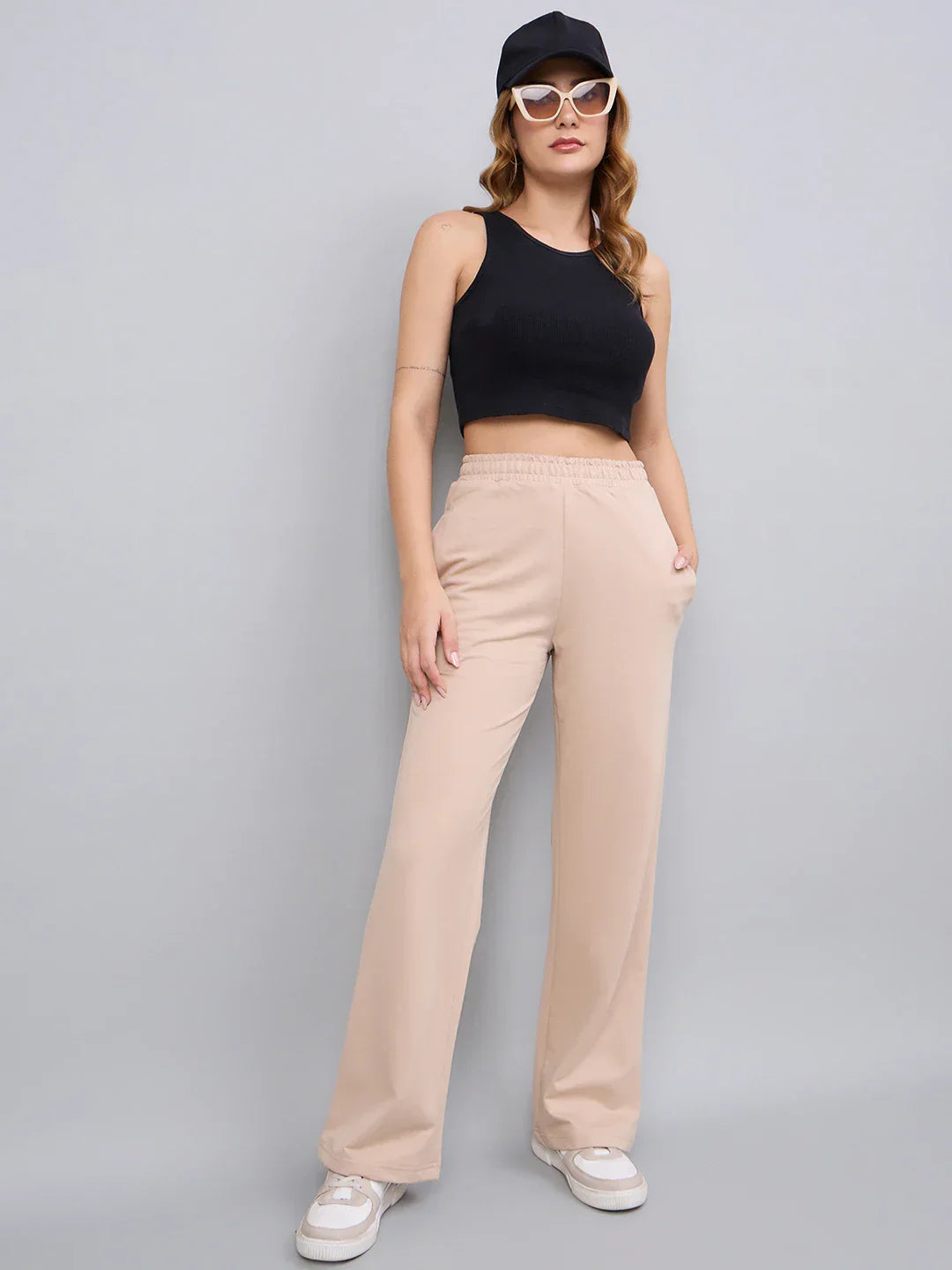 Woman in a black crop top, 98degreenorth beige cotton slim fit trackpants, white sneakers, sunglasses, and black cap.