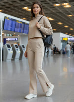 A woman in 98degreenorth beige flared bootcut sweatpants stands at an airport, carrying a black backpack and smiling.