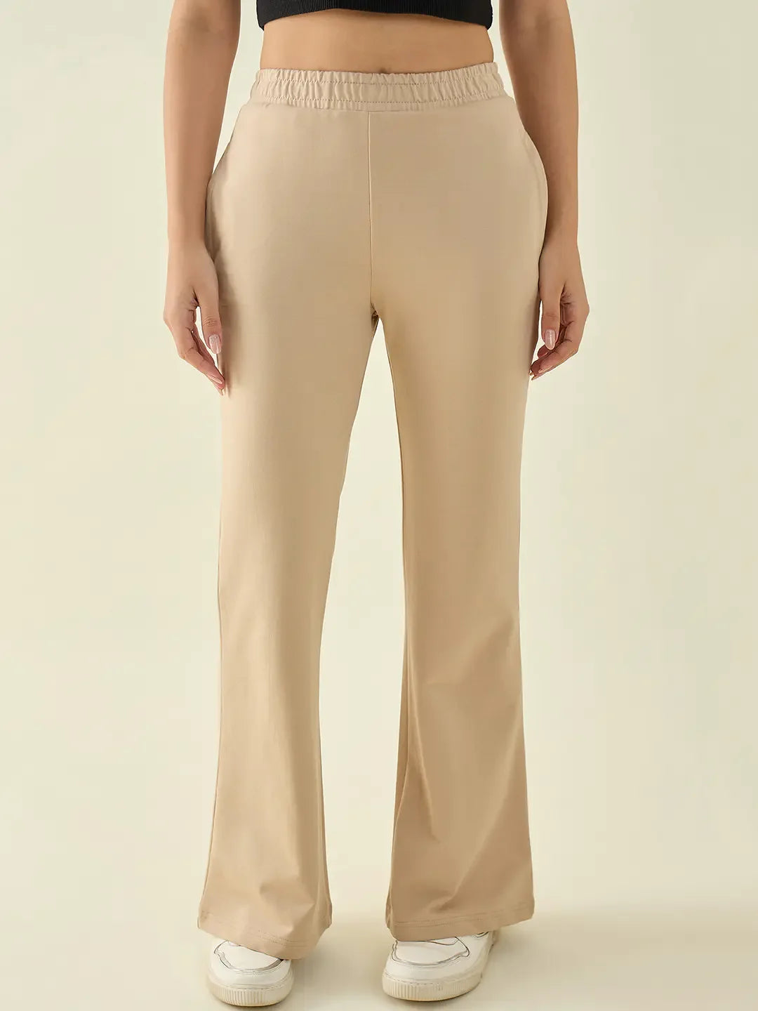 Person wears 98degreenorth Women’s Beige Flared Fit Bootcut Trousers, a black top, and white shoes on a plain light background.