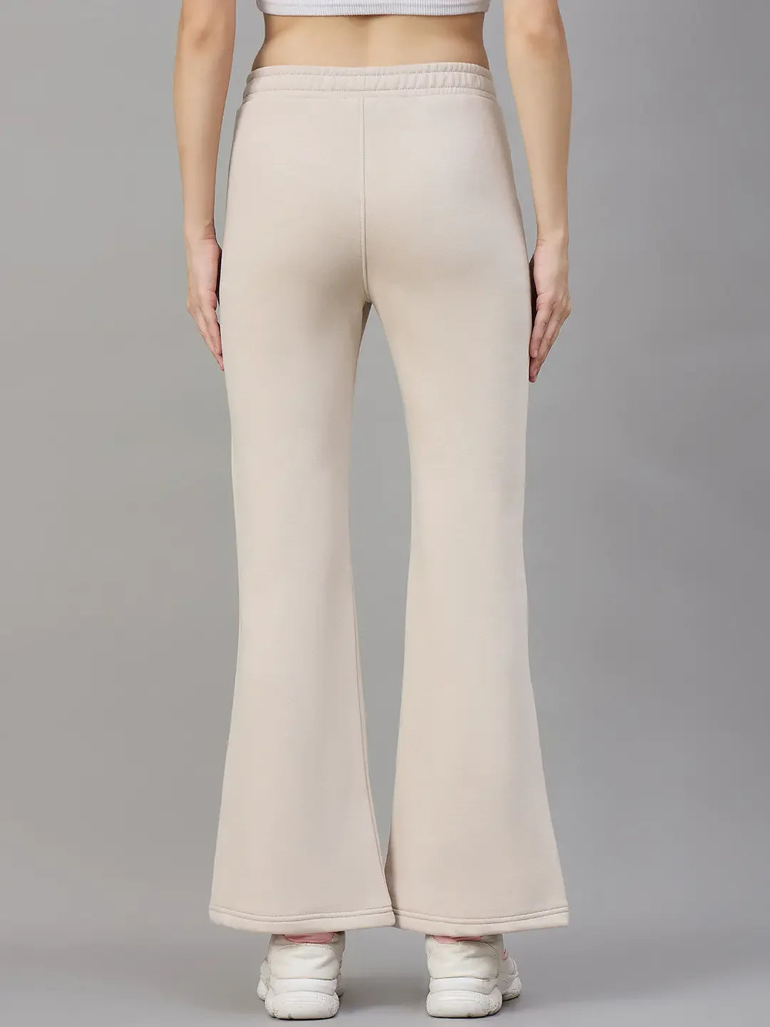 Model in 98degreenorth Women’s Beige Fleece Flared Lounge Pants, shown from the back with white sneakers against gray.