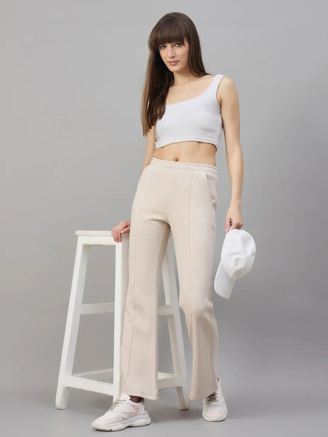 Model in 98degreenorth’s Women’s Beige Fleece Flared Lounge Pants poses with a white cap against a gray background.