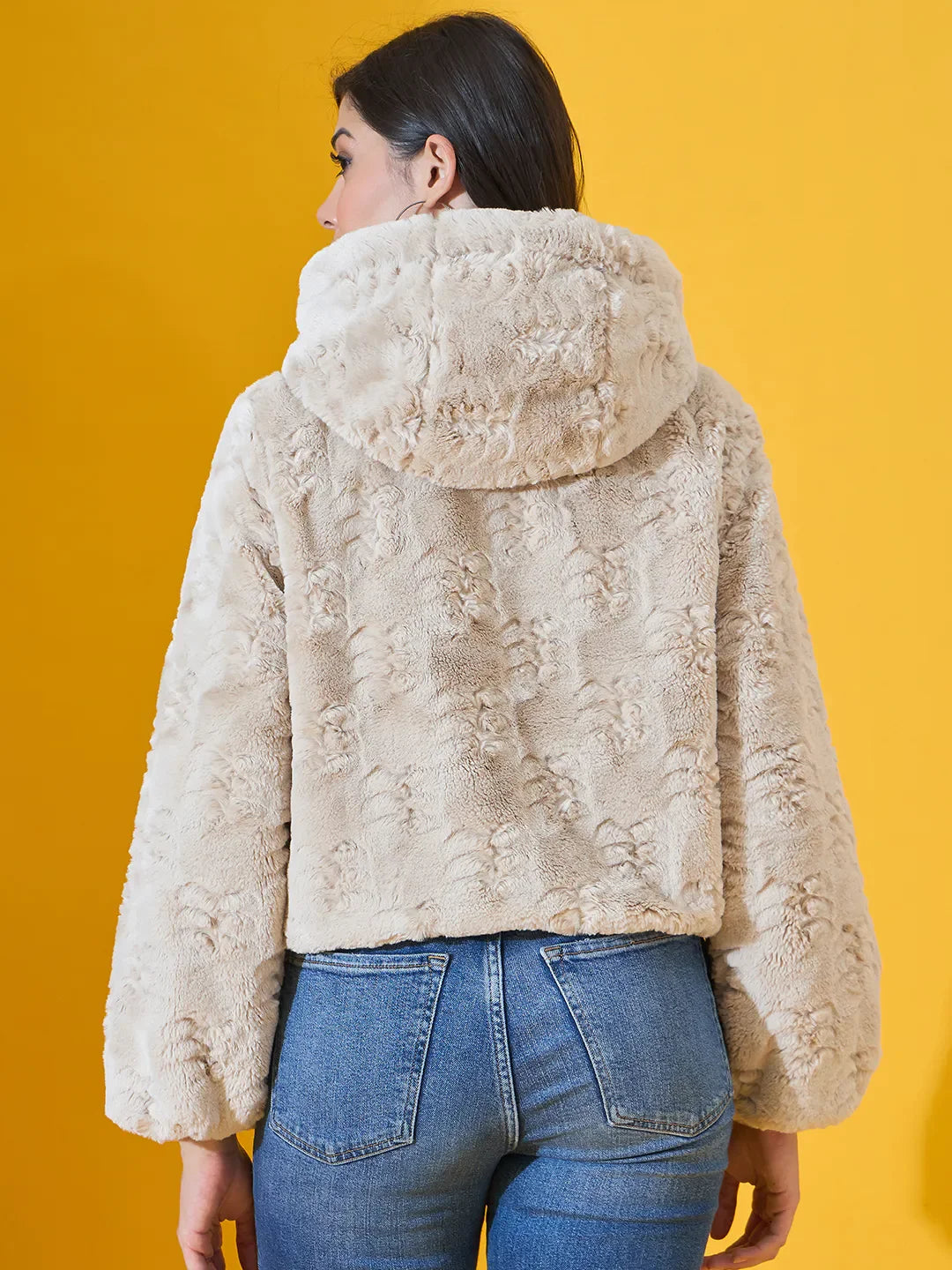 Woman in a 98degreenorth Beige Luxe Faux Fur Hooded Jacket, crop fit winter wear, facing away on yellow background.