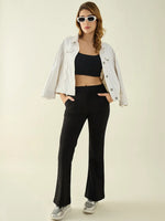 Woman in sunglasses, white jacket, black crop top, and 98degreenorth black flared bootcut stretch trousers stands hands in pockets.