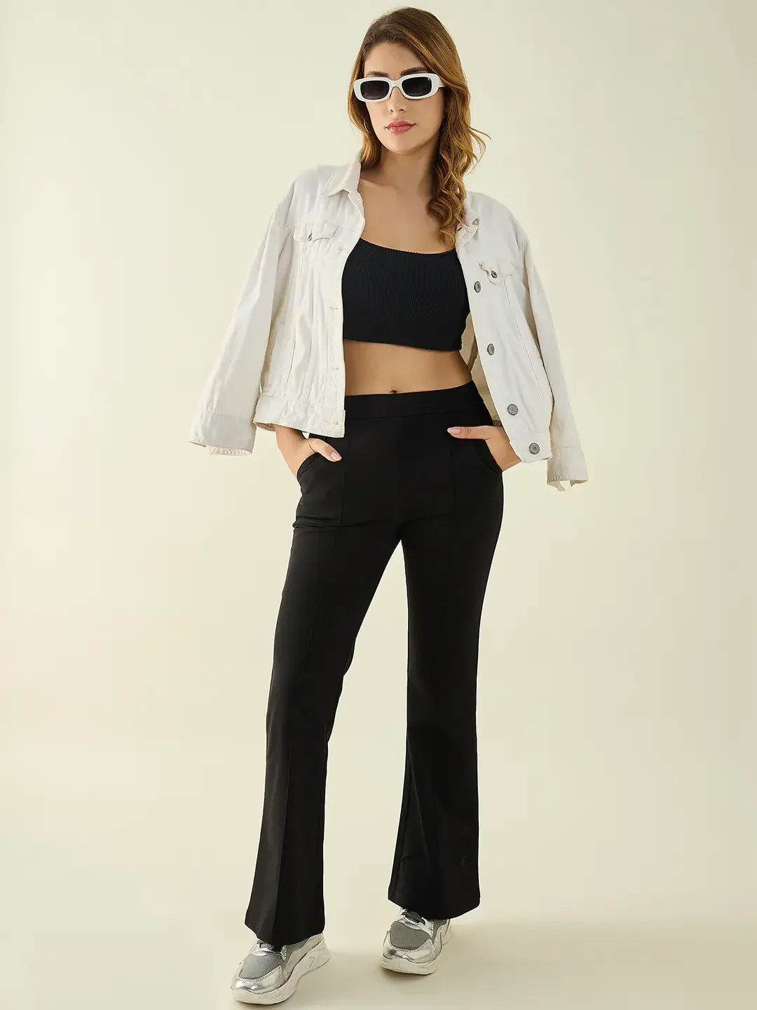 Woman in sunglasses, white jacket, black crop top, and 98degreenorth black flared bootcut stretch trousers stands hands in pockets.