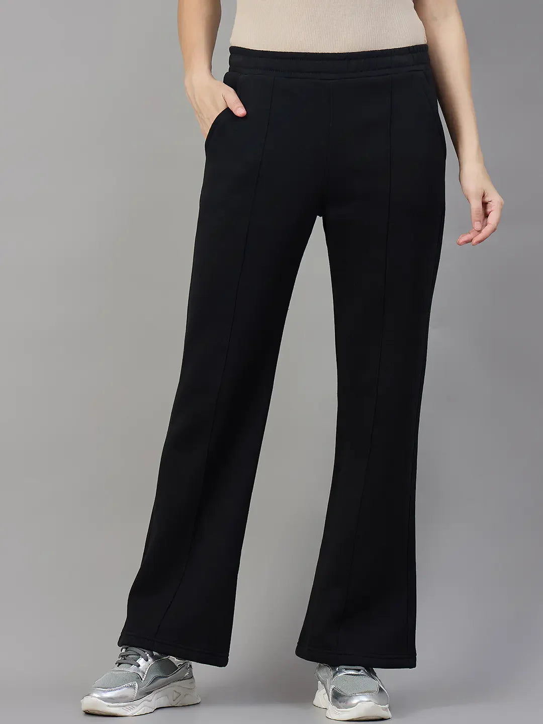 Model in 98degreenorth Women’s Black Fleece Flared Sweatpants, beige top, silver sneakers; hand in pocket, gray backdrop.