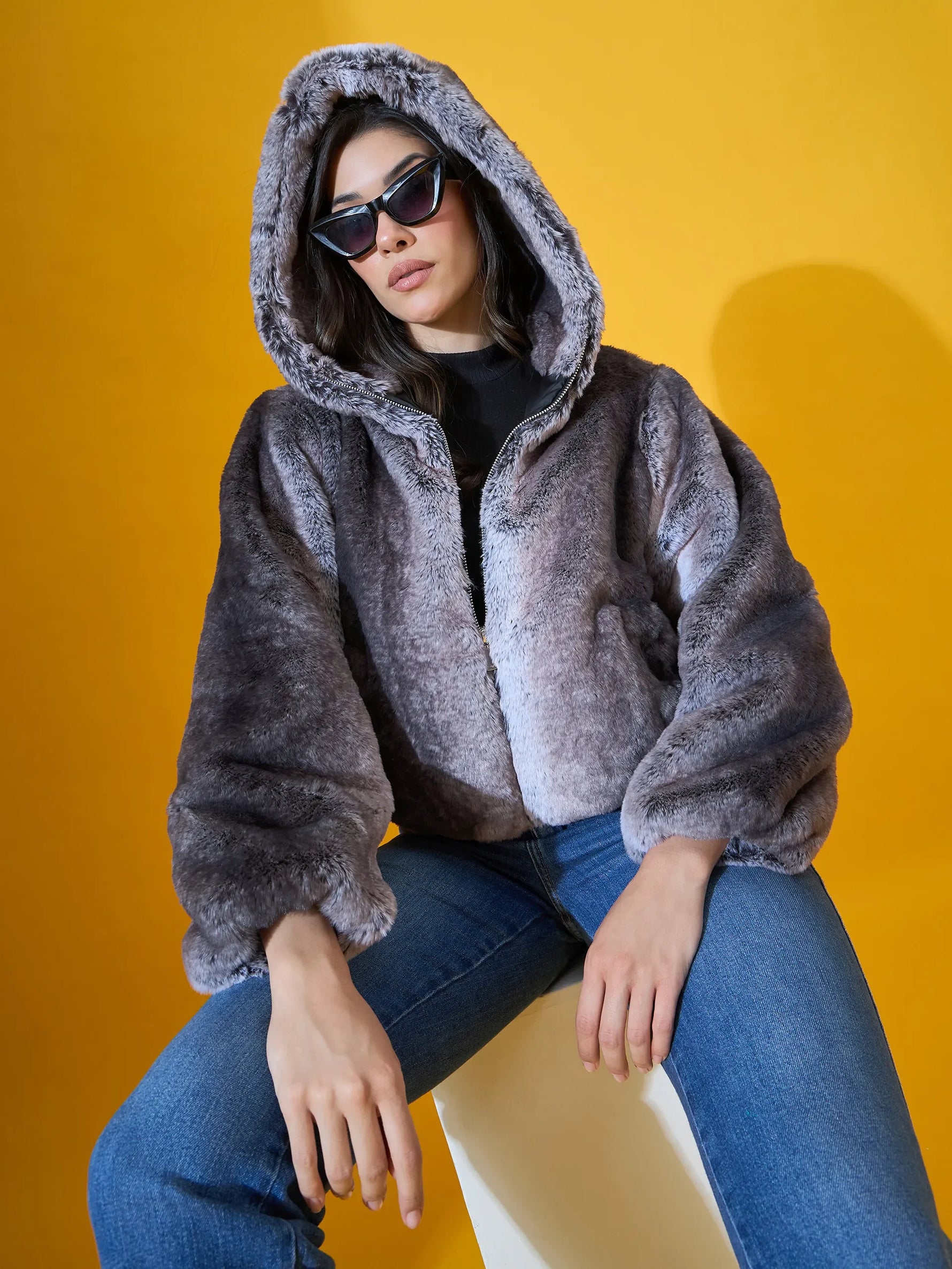 Woman wears the 98degreenorth Women's Black Premium Faux Fur Jacket with plush hood, seated against a yellow background.