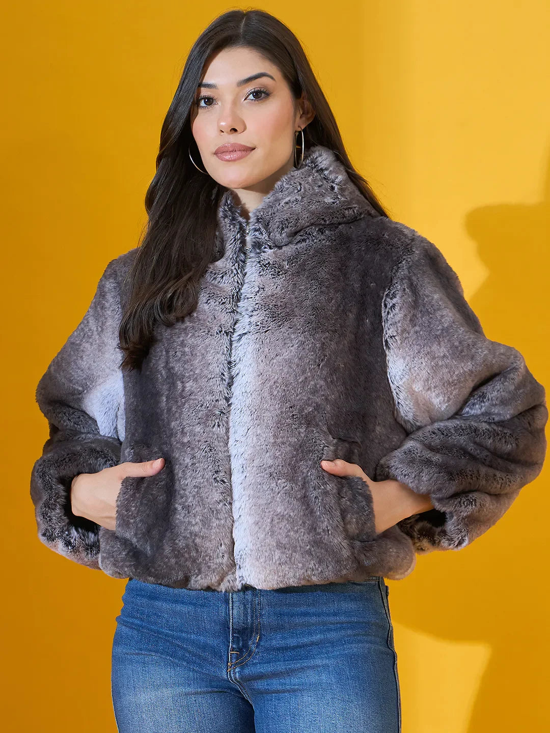 Woman in a 98degreenorth Women’s Black Premium Faux Fur Jacket with hood and blue jeans stands by a yellow background.