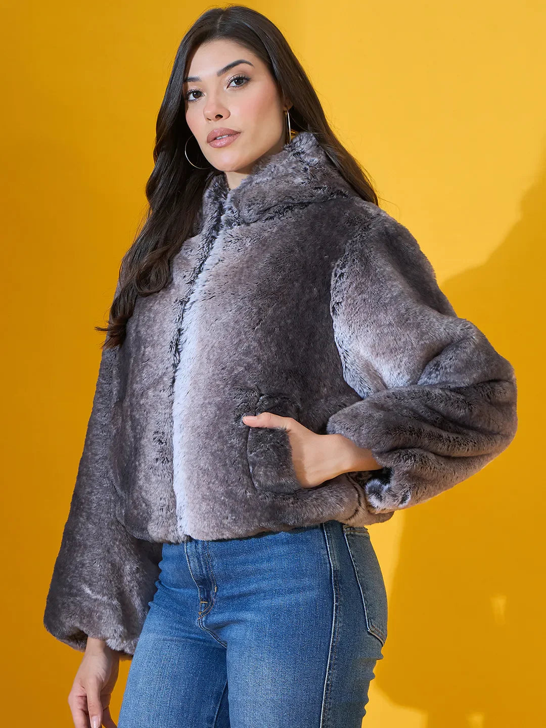 Woman in a 98degreenorth Women’s Black Premium Faux Fur Jacket with plush hood poses against a yellow background.