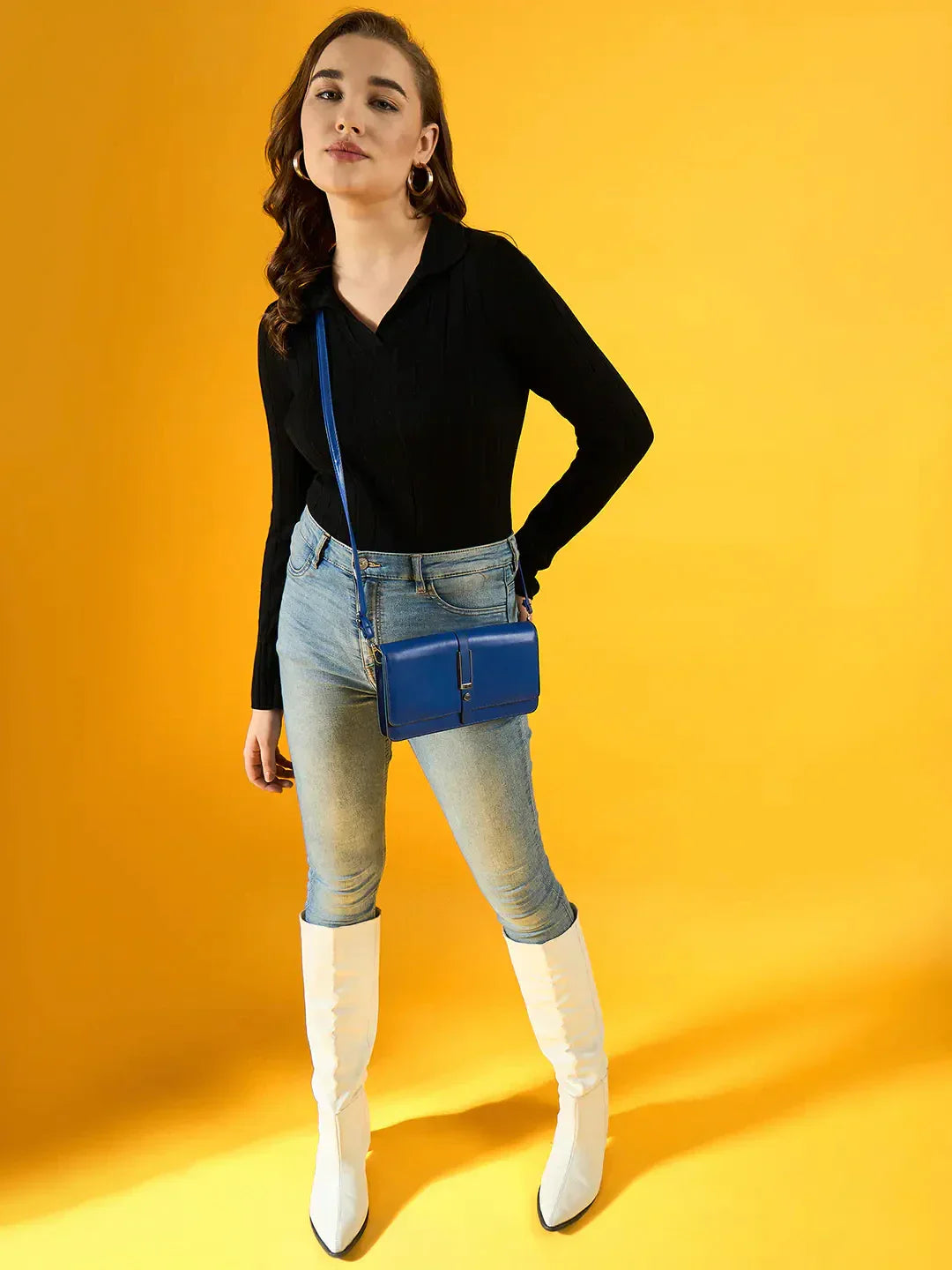 Woman in 98degreenorth Black Ribbed Collar Neck Sweater, blue jeans, white boots & blue purse against yellow background.