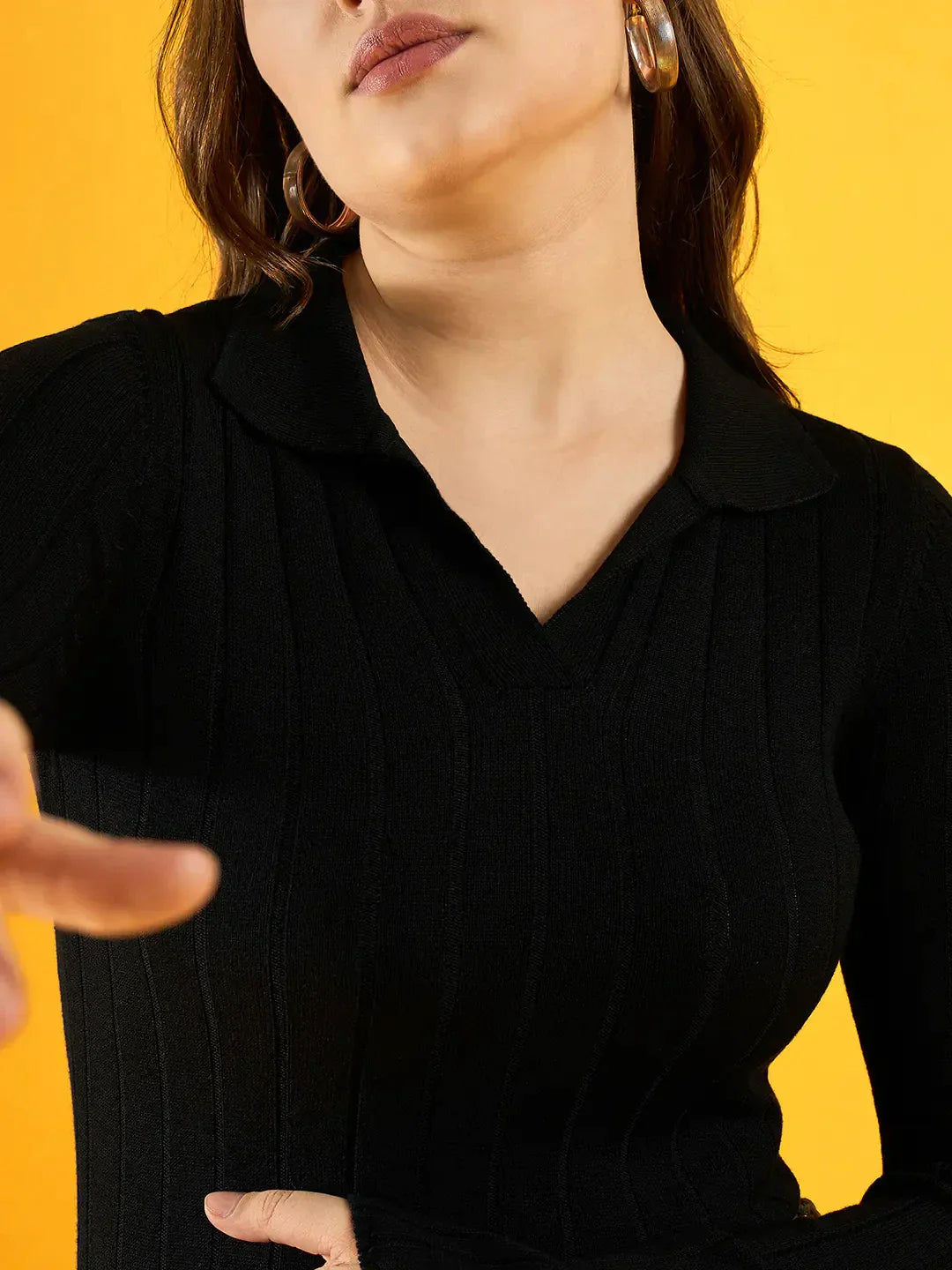 Woman wears a 98degreenorth Women’s Black Ribbed Collar Neck Sweater, posing in hoop earrings on a yellow background.