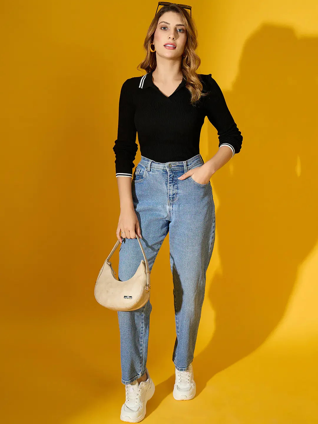 Woman in 98degreenorth’s black ribbed collared knit sweater and blue jeans, holding a beige handbag against yellow.