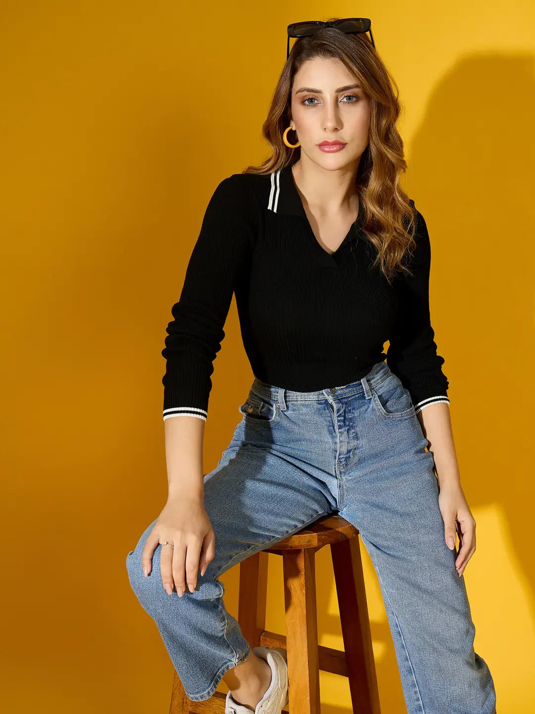 Woman wears 98degreenorth’s black ribbed collared knit sweater and blue jeans, seated on a stool with yellow backdrop.