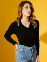 A woman wears the 98degreenorth Women’s Black Ribbed Collared Knit Sweater with blue jeans against a yellow background.