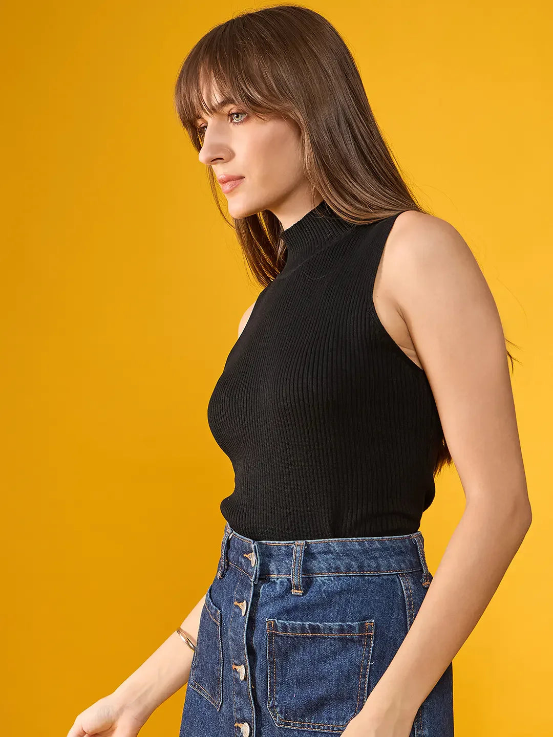 Woman in a 98degreenorth black ribbed high neck sleeveless knit top and denim skirt stands against a yellow background.