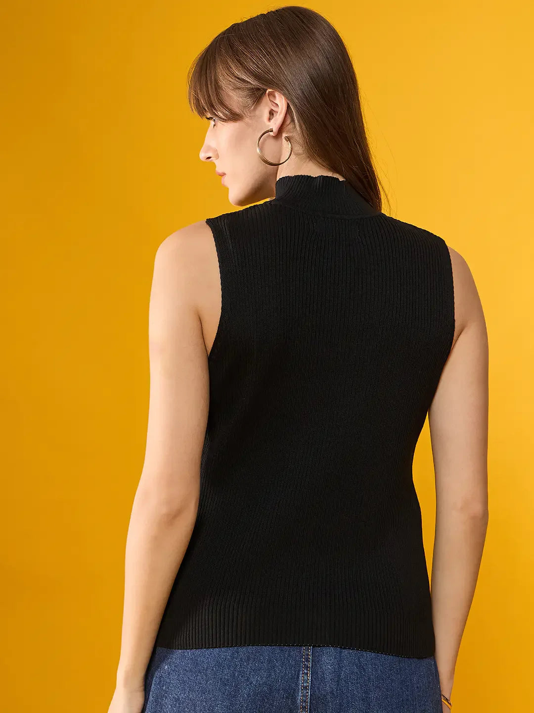 Woman in a 98degreenorth black ribbed high neck sleeveless slim fit knit top, paired with blue jeans on a yellow background.