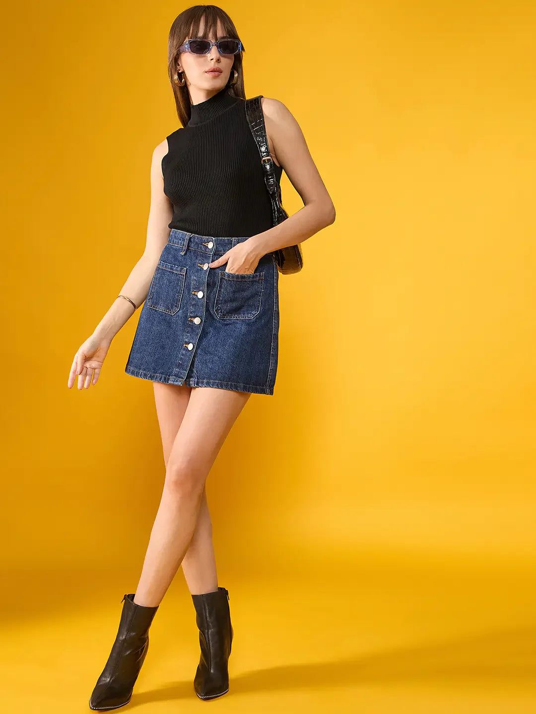 Woman wears 98degreenorth's black ribbed high neck slim fit tank with a denim skirt, boots, shades, and bag on yellow.