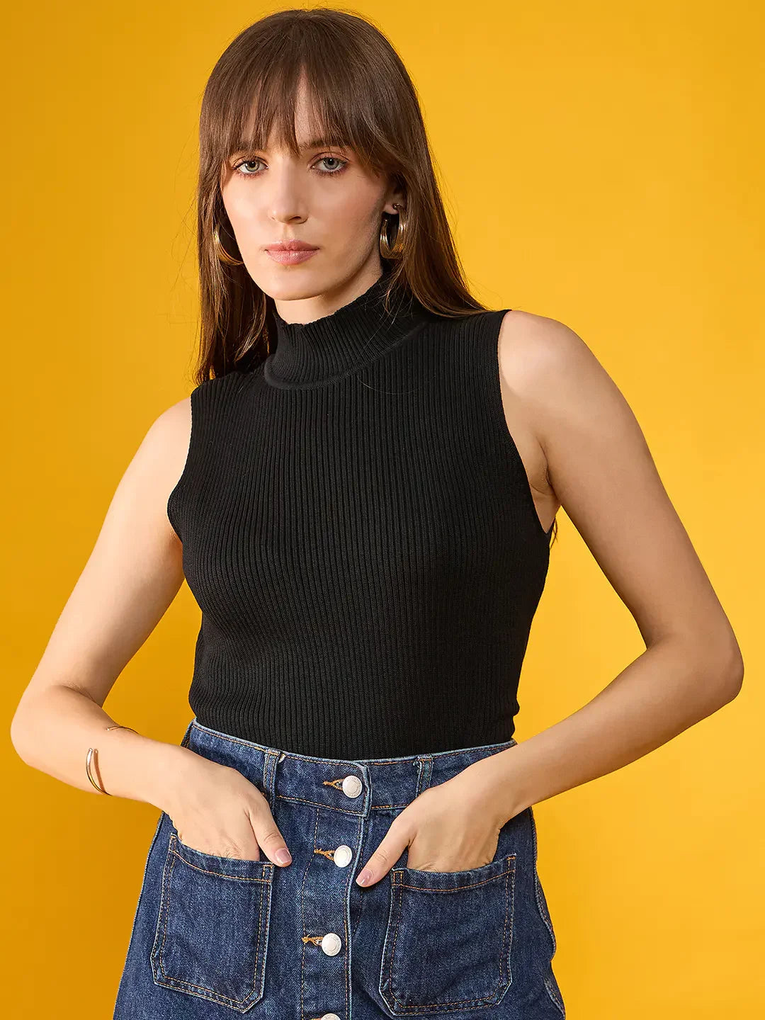 Woman wears 98degreenorth’s black ribbed high neck sleeveless knit top with a blue skirt, posing by a yellow wall.