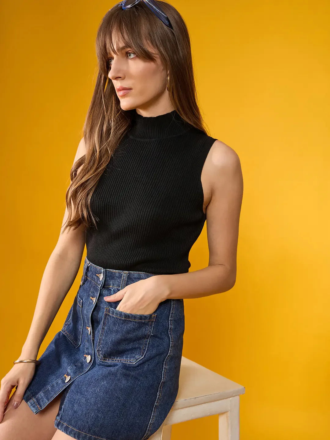 Woman wears 98degreenorth Black Ribbed High Neck Sleeveless Knit Top, paired with a denim skirt against yellow backdrop.