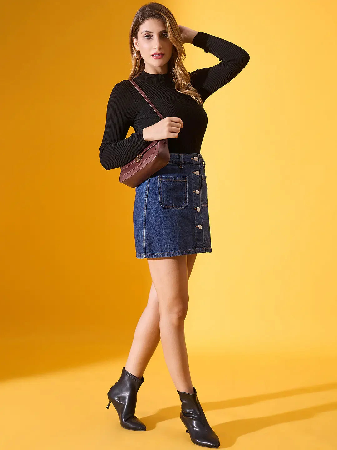 Woman poses against yellow in a 98degreenorth black ribbed high-neck slim fit sweater, denim skirt, boots, and brown bag.
