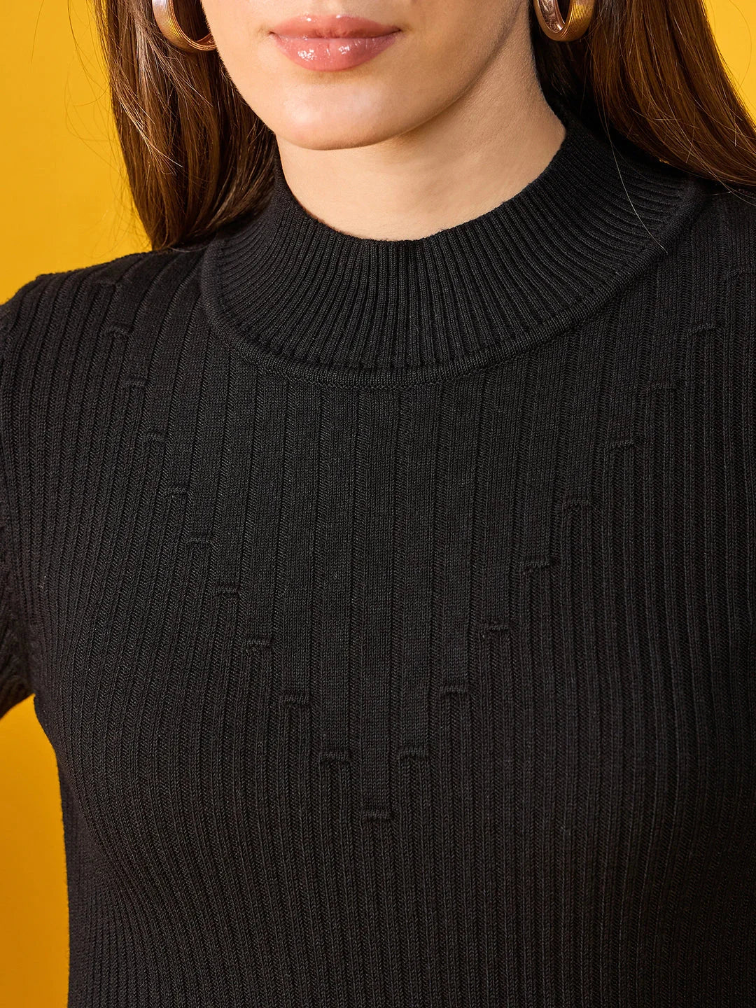 A woman models a 98degreenorth black ribbed slim fit turtleneck sweater and gold hoops against a yellow background.