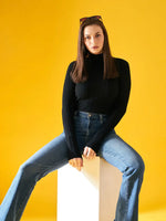 Woman wears 98degreenorth Black Ribbed Turtleneck Sweater, paired with blue jeans and sunglasses, on a yellow background.