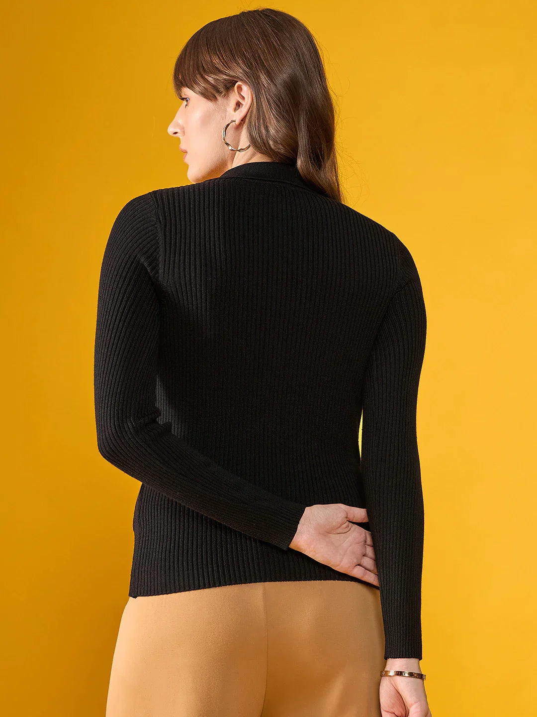 A woman wears a 98degreenorth Women’s Black Ribbed Sweater with tan pants, facing away against a yellow background.