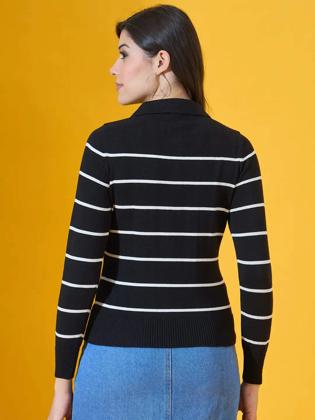Model in 98degreenorth Women’s Black Striped Collared Knit Sweater and jeans, posed before a yellow backdrop.