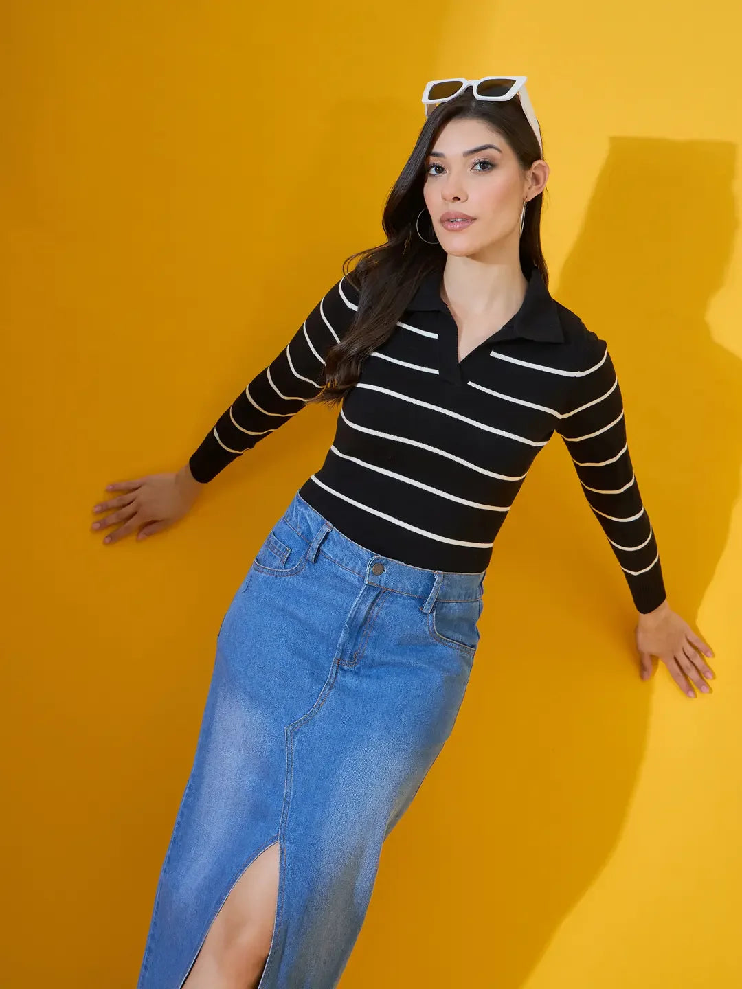 Woman models 98degreenorth’s black striped collared knit sweater with a denim skirt and white sunglasses, yellow background.