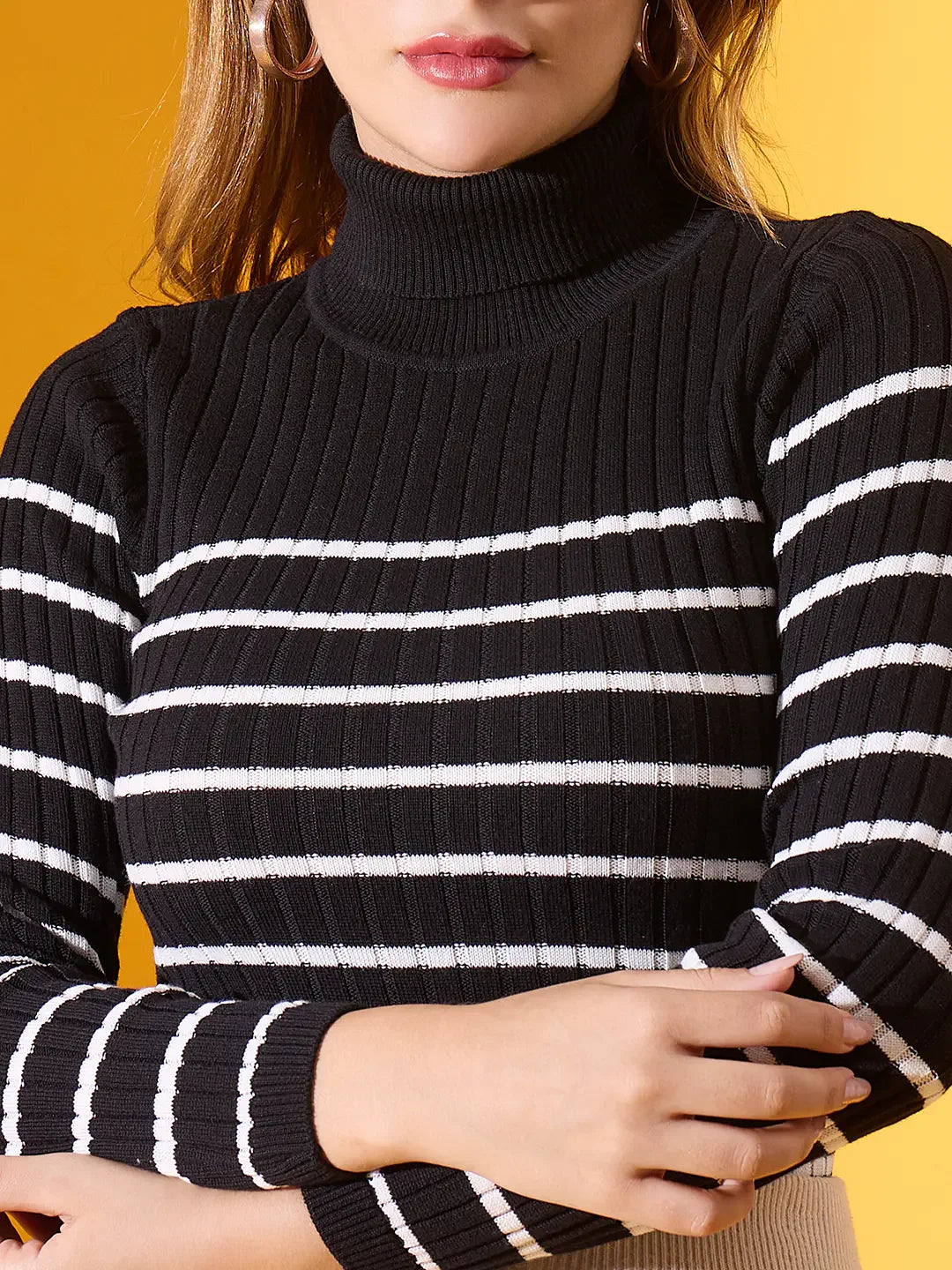 Woman in 98degreenorth Women’s Black Striped Ribbed High Neck Sweater, arms crossed, on a yellow background.
