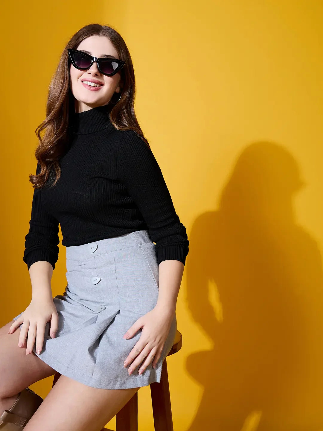 A woman in sunglasses and a 98degreenorth Women’s Black Turtle-Neck Ribbed Sweater sits on a stool by a yellow background.