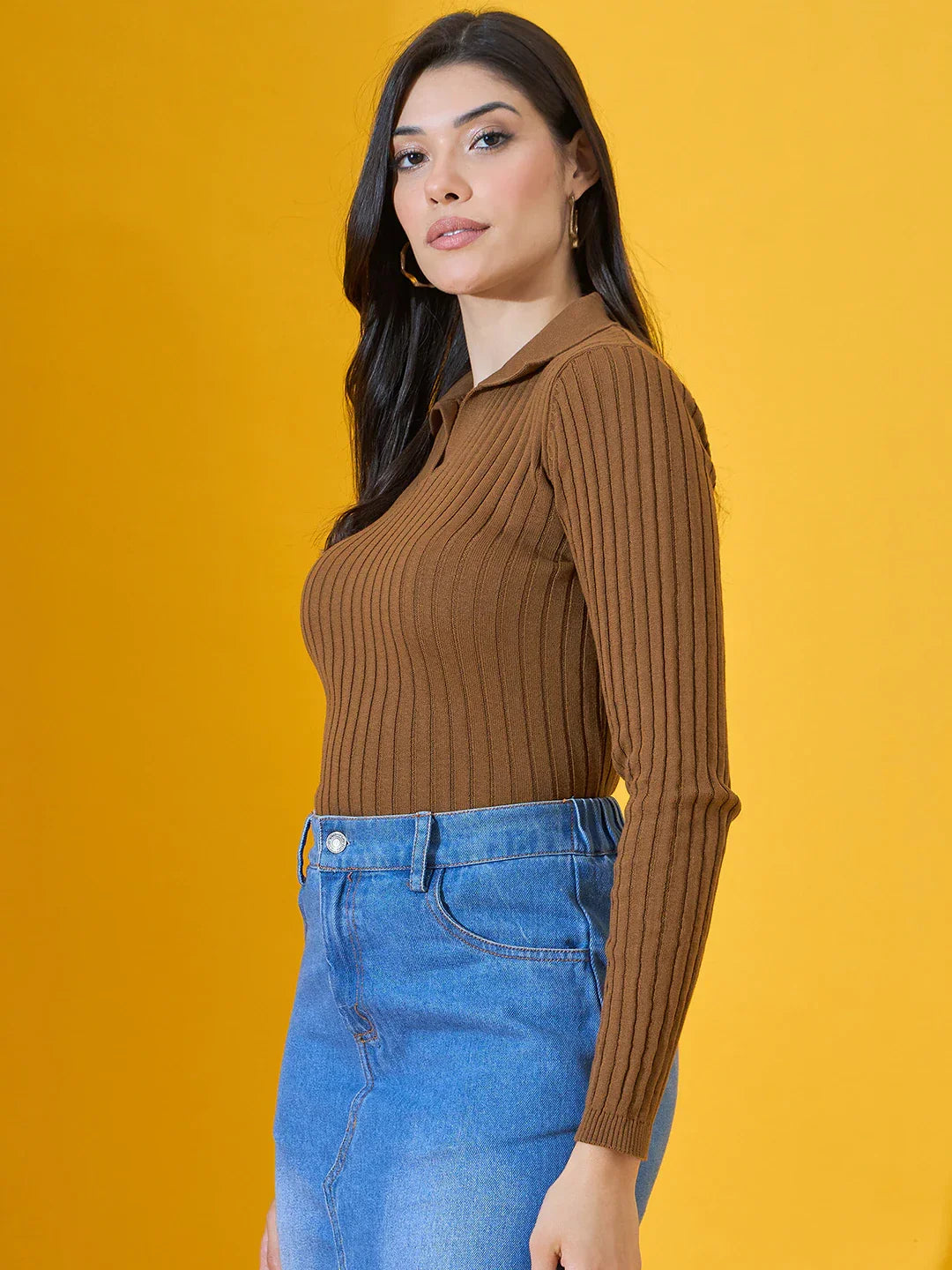 Woman in a 98degreenorth brown ribbed collar neck slim fit sweater and blue jeans stands before a yellow background.