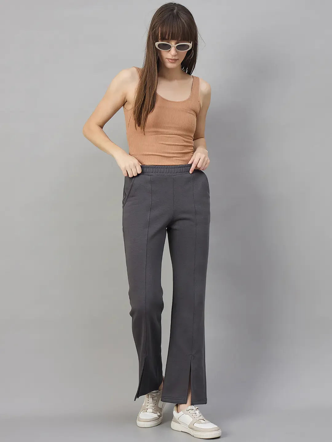 Woman in a tan tank and 98degreenorth charcoal grey bootcut sweatpants with side slits stands against a gray background.