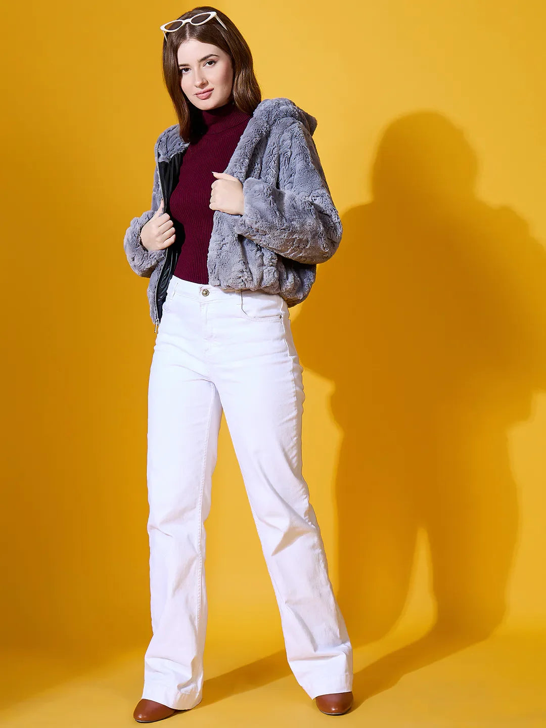 Woman models 98degreenorth's charcoal grey faux fur jacket, maroon top, white pants, and brown shoes against yellow.
