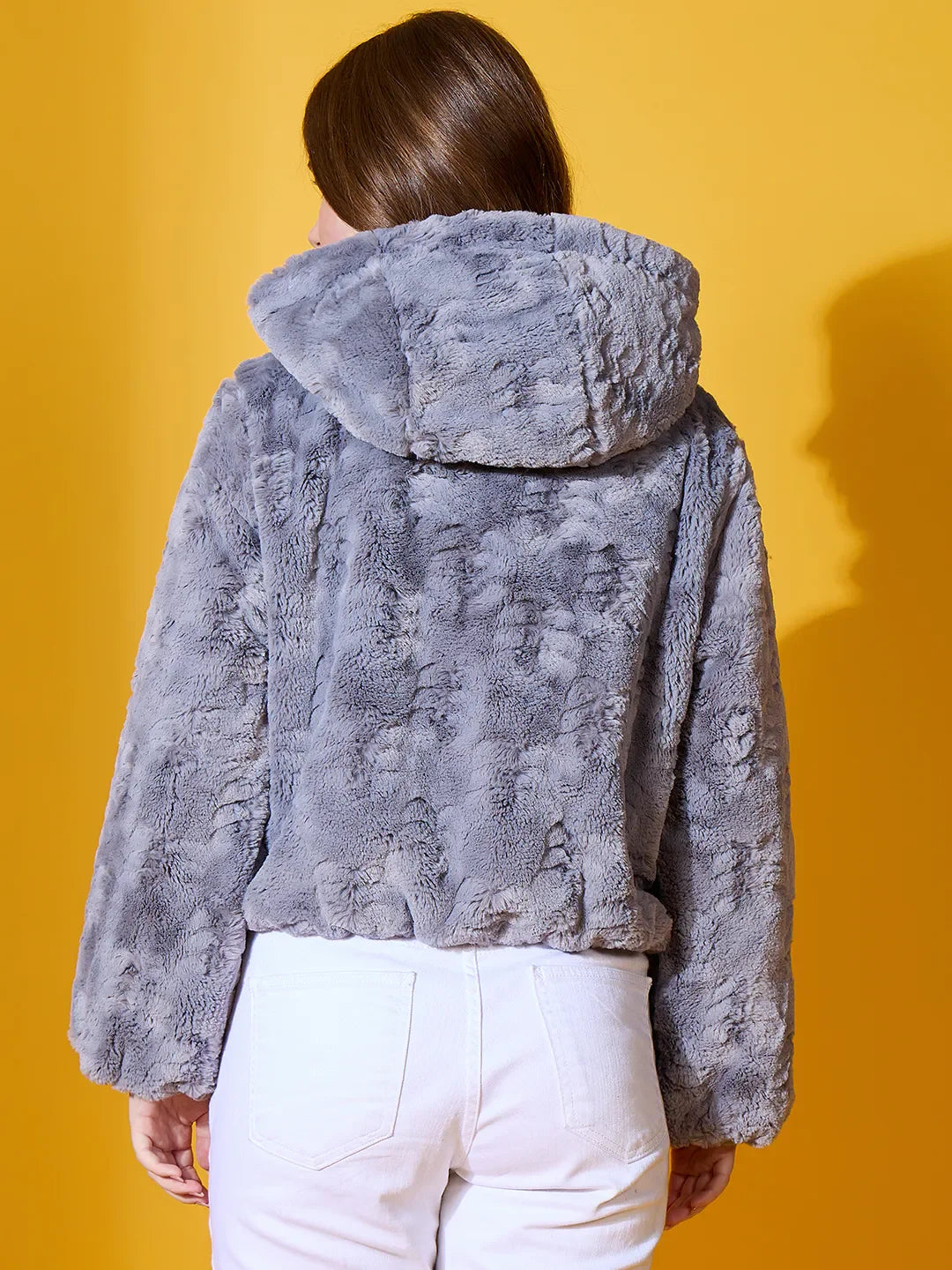 A woman wears the 98degreenorth Women’s Charcoal Grey Faux Fur Jacket, standing before a bold yellow background.