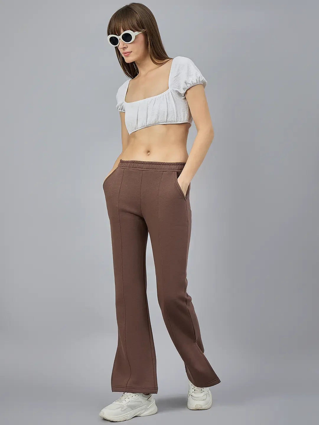 Woman in white crop top and 98degreenorth cocoa brown flared sweatpants stands in white sneakers and sunglasses.