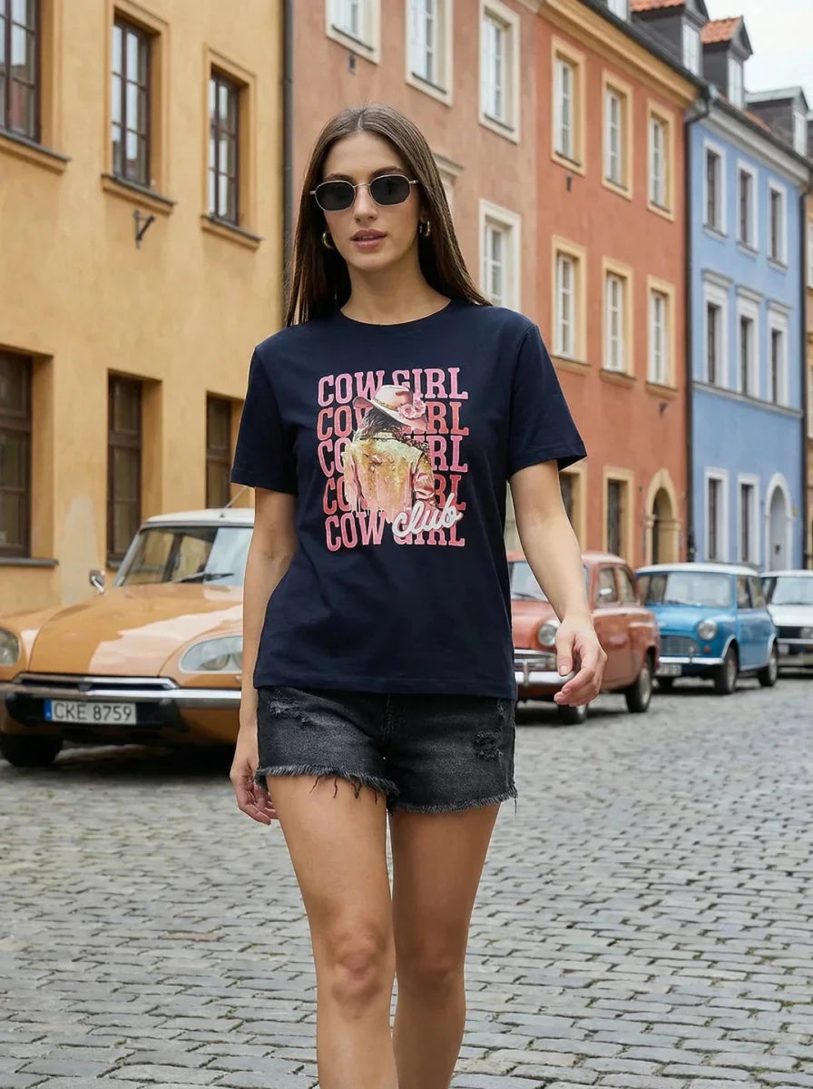 A woman in sunglasses wears the 98degreenorth Women’s Cotton Typography Graphic Tee on a vibrant cobblestone street.