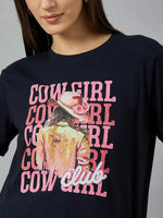 A woman wears a 98degreenorth Women’s Cotton Typography Graphic Tee featuring “COWGIRL club” and a pink-hat cowgirl.